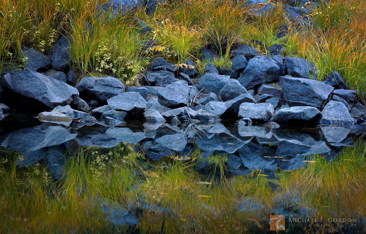 Autumn-hued grasses and shrubs and blue granite reflected in the tranquil Merced River.