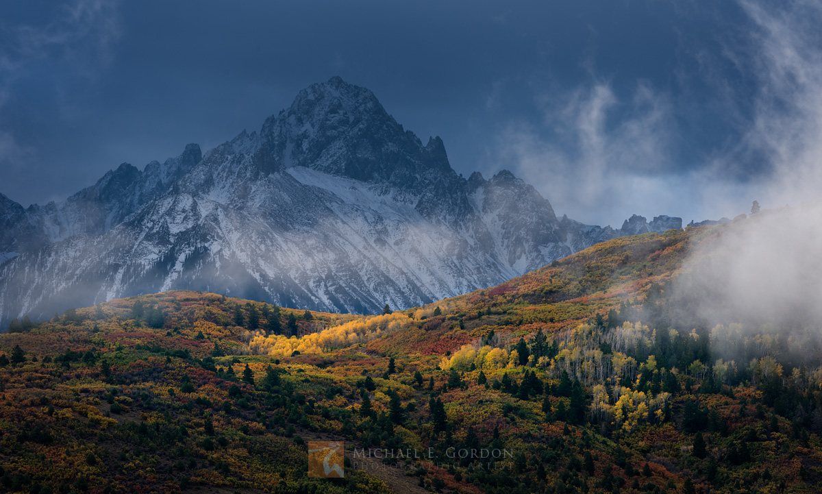 A momentary break in the clouds spotlights a colorful autumn grove of Quaking Aspen (Populus tremuloides), Gambel's Oak (Quercus...